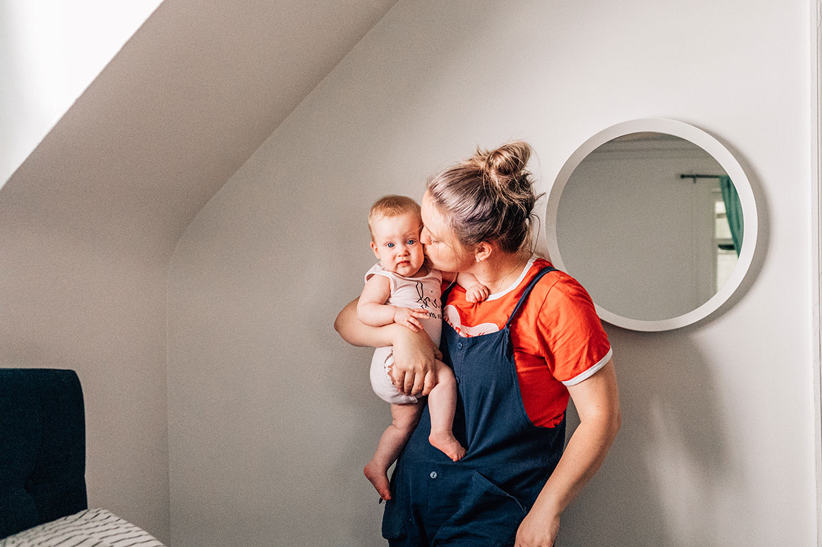 a mama wearing a red shirt and navy overalls kisses her baby on the cheek.