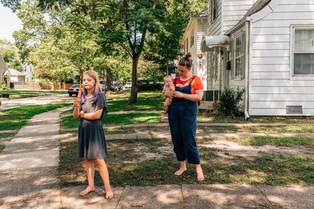 a mother and her children stand outside their home along the sidewalk eating orange popsicles