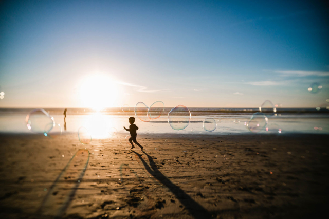 A silhouette of a little boy running down the beach at sunset chasing bubbles.