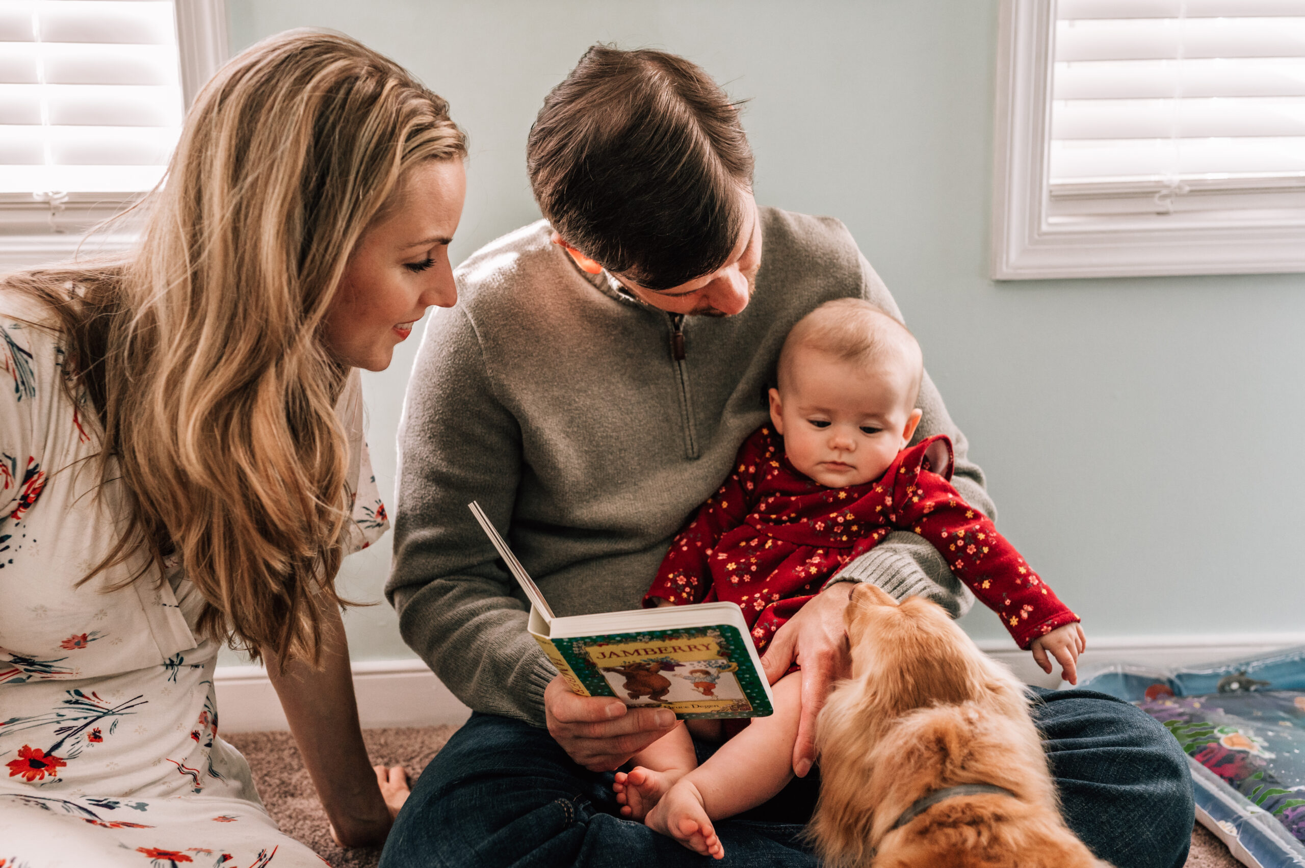 A mom and dad sit reading to their baby while the family dog checks on baby.