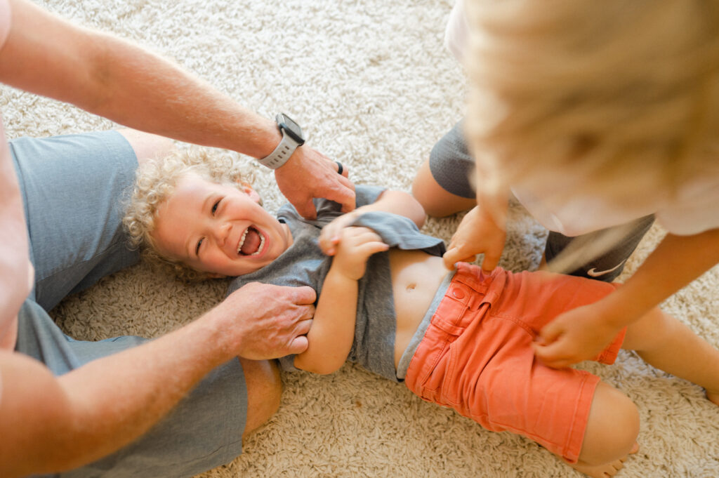 Little boy laughs as dad and brother tickle him during in-home family photo session.