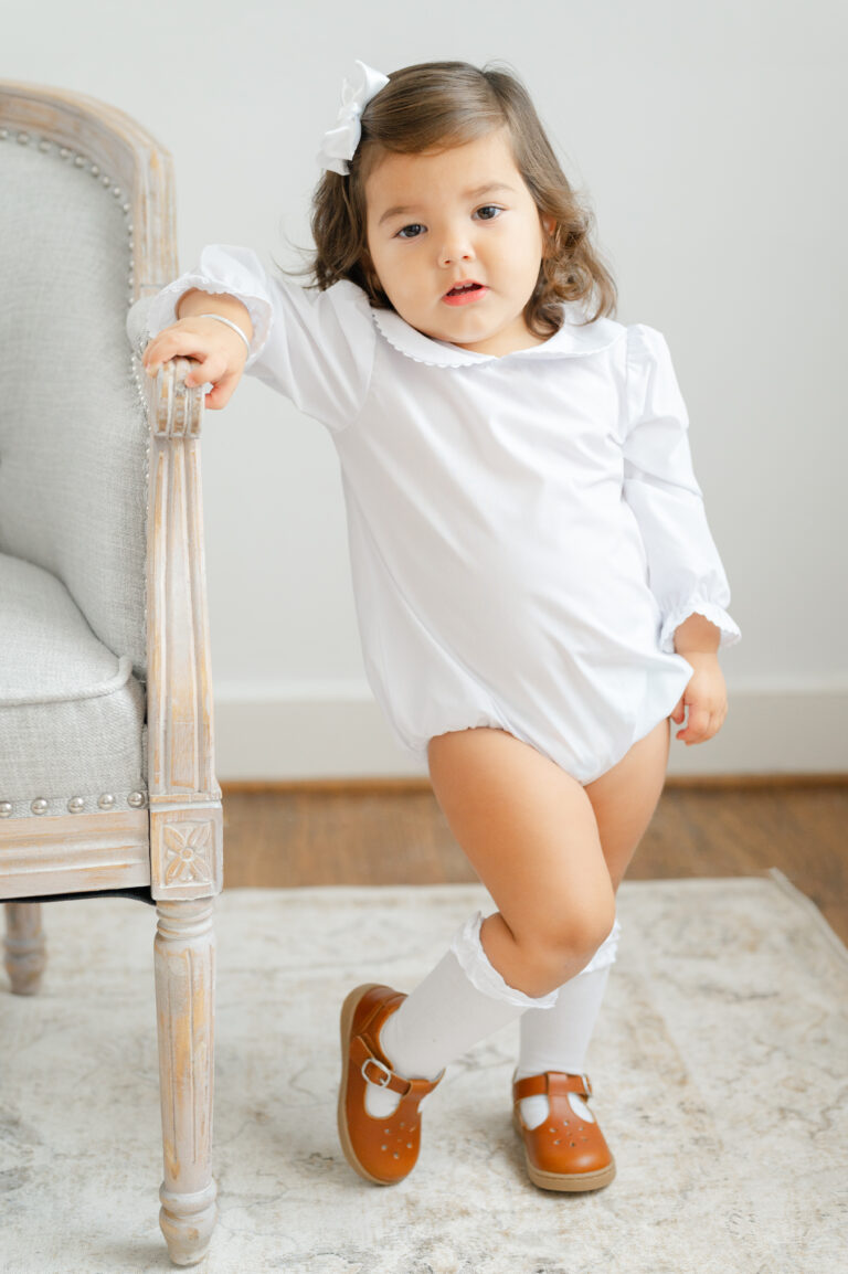 Little girl in classic clothing poses next to a tufted couch during heirloom photography session.