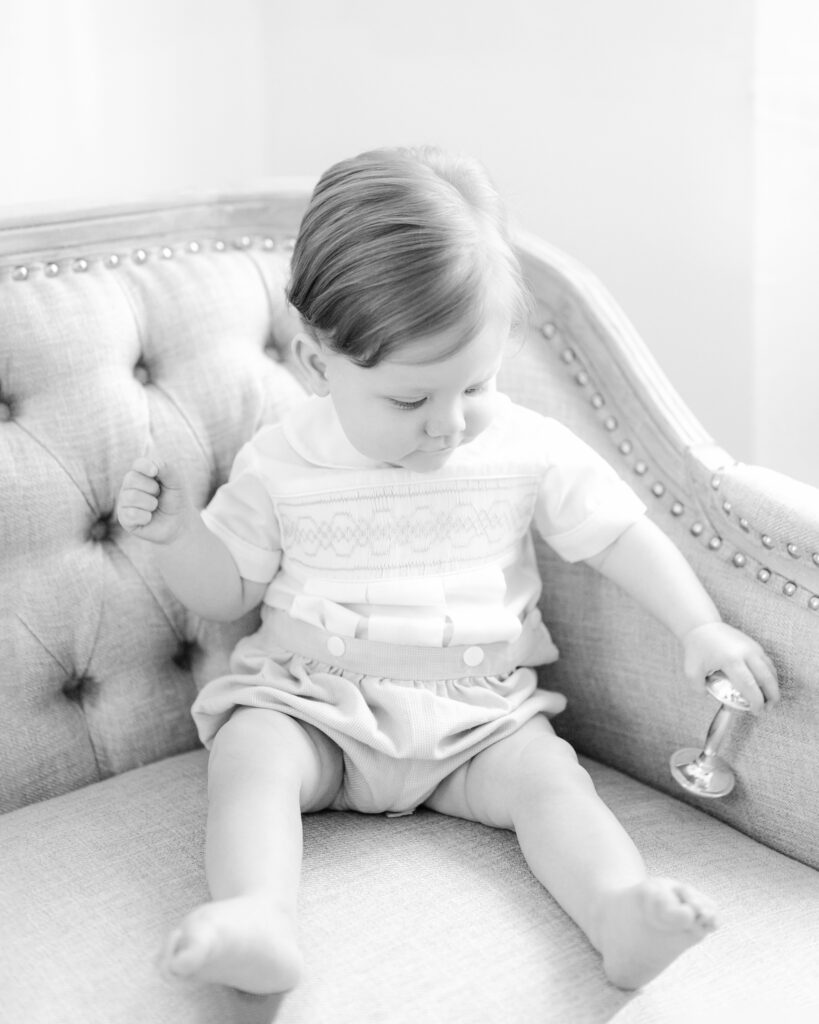 Little boy wearing classic smocked outfit holds on to silver rattle.
